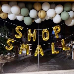 storefront window with a balloon garland of green, gold, and white balloons and below gold letter balloons spelling out shop small