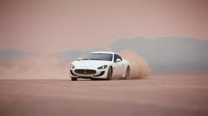 The image shows a white Maserati GT drifting sideways in a dusty desert landscape.
