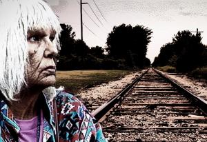 A weathered older woman stares into space in the foreground. Railroad tracks in the background.